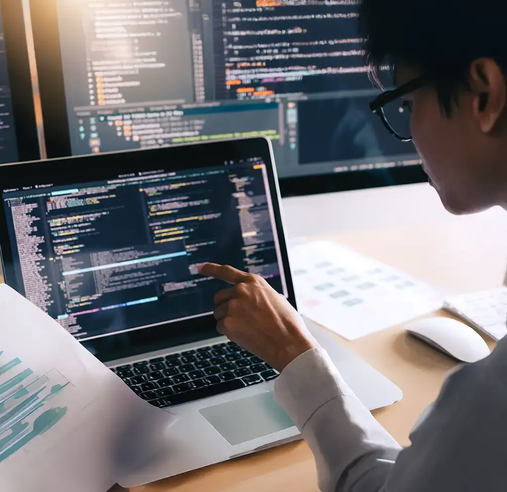Developer reviewing and debugging code on a laptop with multiple monitors in a workspace.