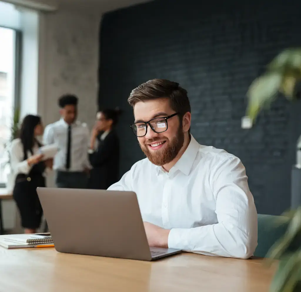Smiling professional working on a laptop at a desk with colleagues conversing in the background.