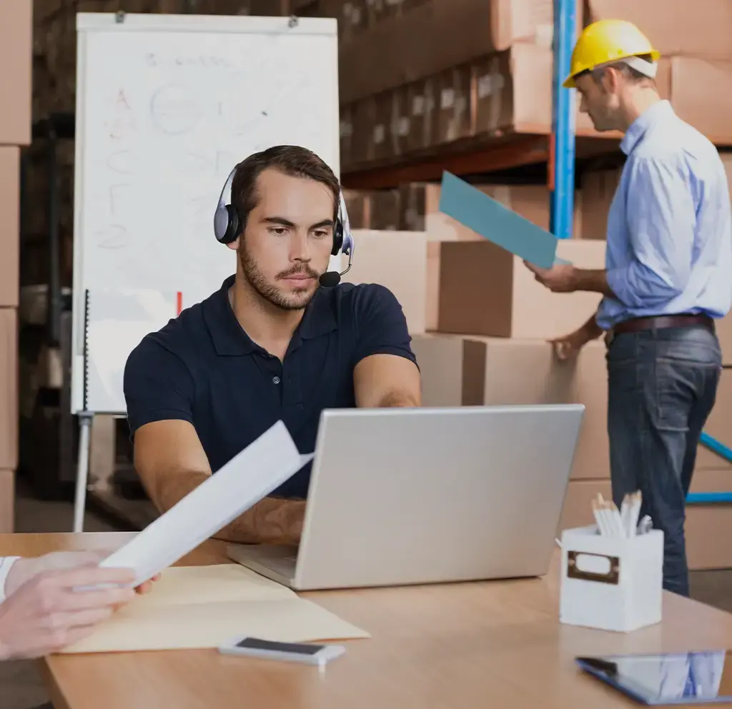 Logistics coordinator with headset manages supply chain operations on laptop in warehouse setting.