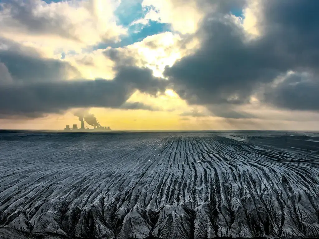 Industrial power plant emitting smoke beyond a vast, barren landscape under dramatic cloud cover.