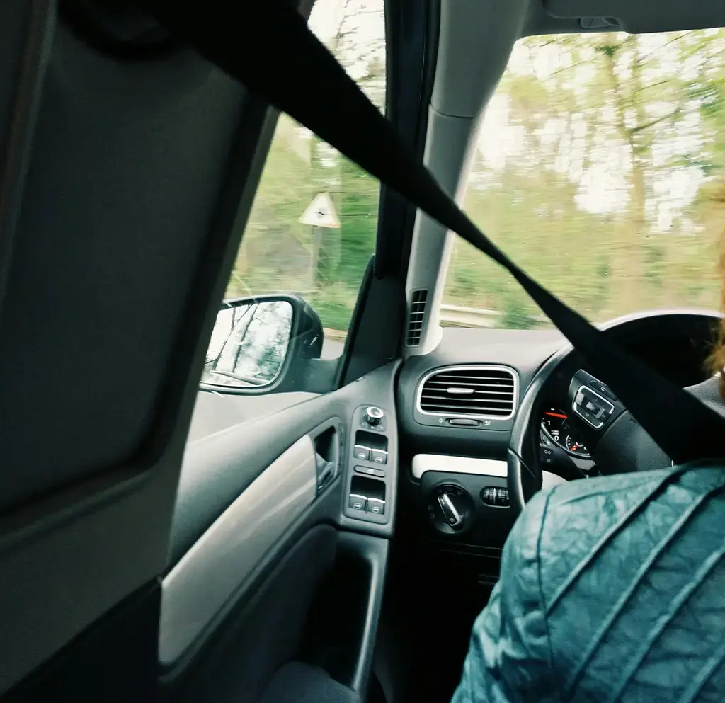 View from a car’s back seat showing dashboard, steering wheel, and driver on a road with trees.