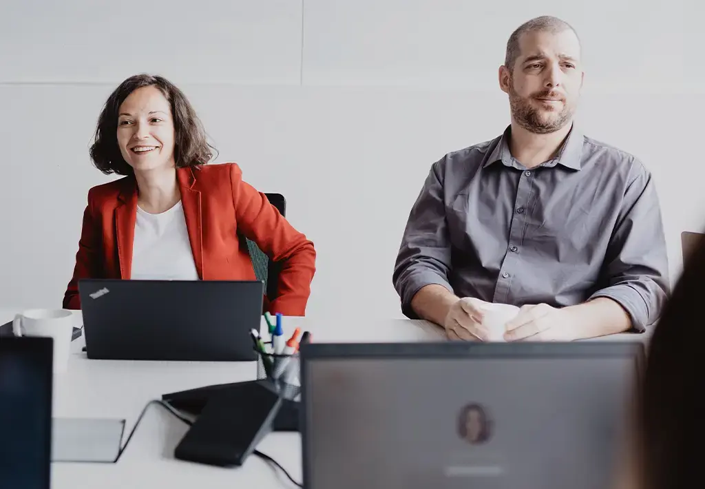 Professionals engaged in a collaborative meeting around a conference table with laptops open.