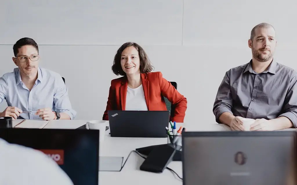 Professionals engaged in a collaborative meeting around a conference table with laptops open.