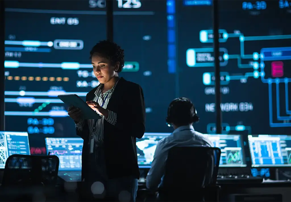 Professional reviewing data on tablet in control room with digital dashboards in background.