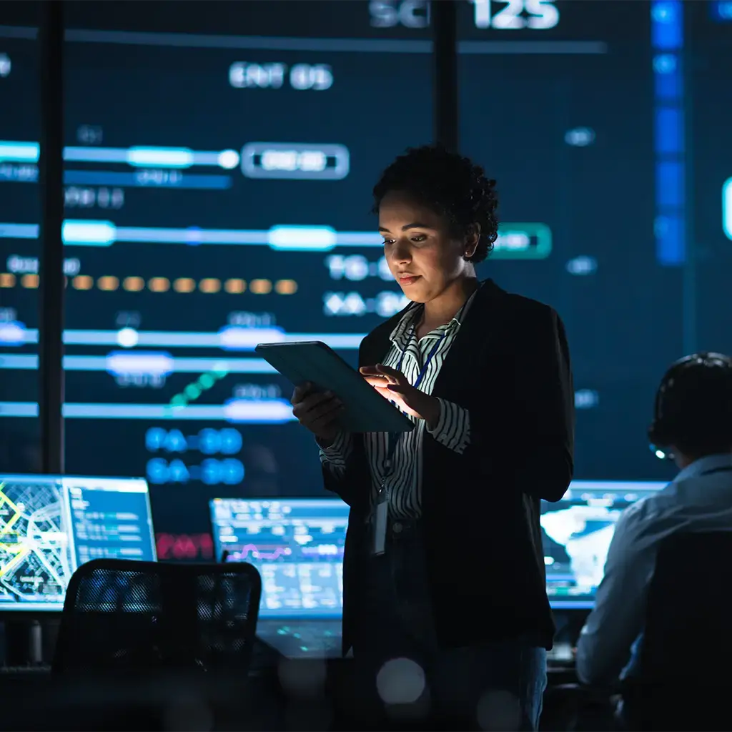 Professional reviewing data on tablet in control room with digital dashboards in background.