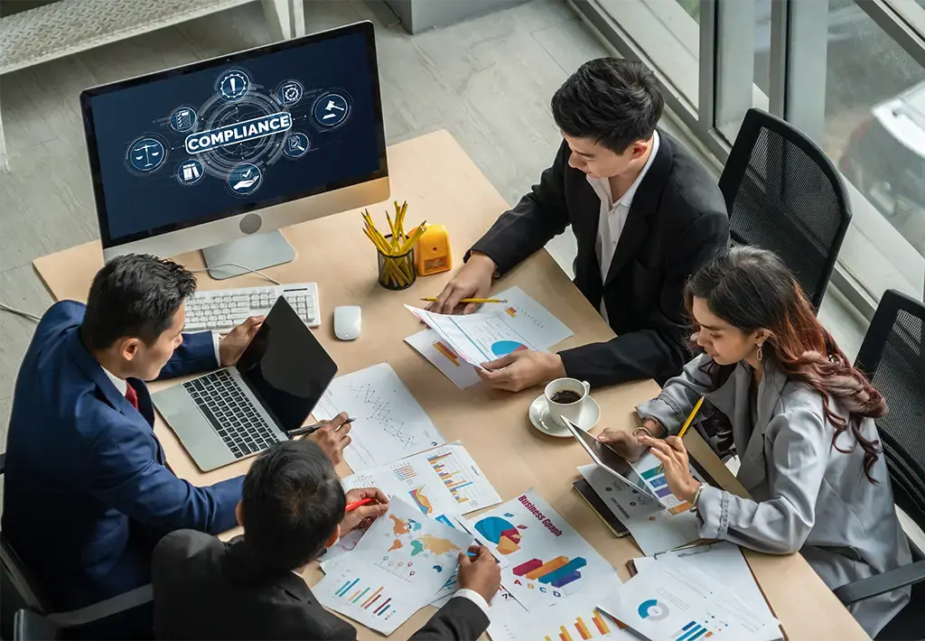 Team collaborating on compliance strategy with charts and digital reports in modern office.