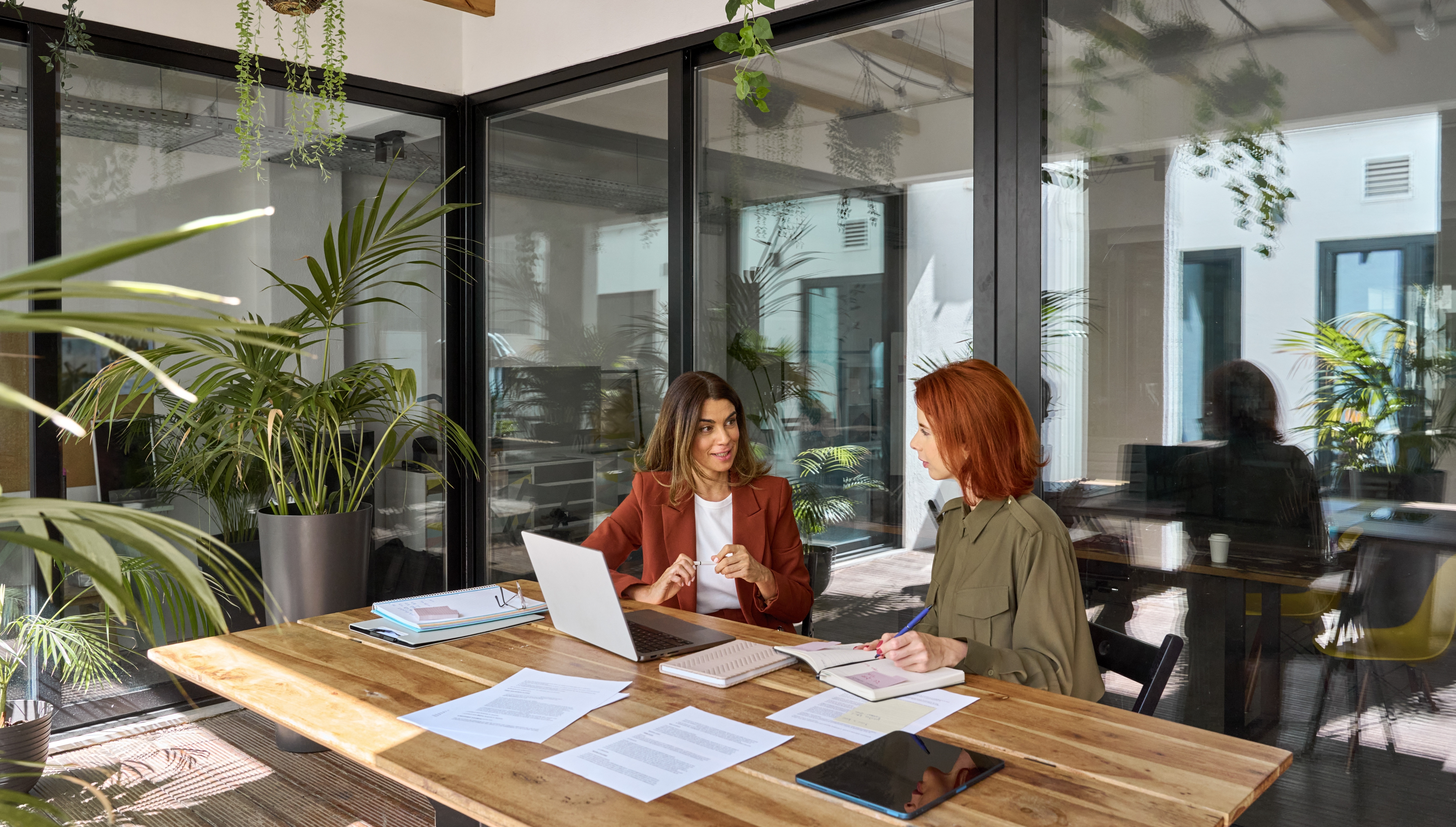 Two women sit at a wooden table in a modern office, discussing work. Papers, a laptop, and plants decorate the space.
