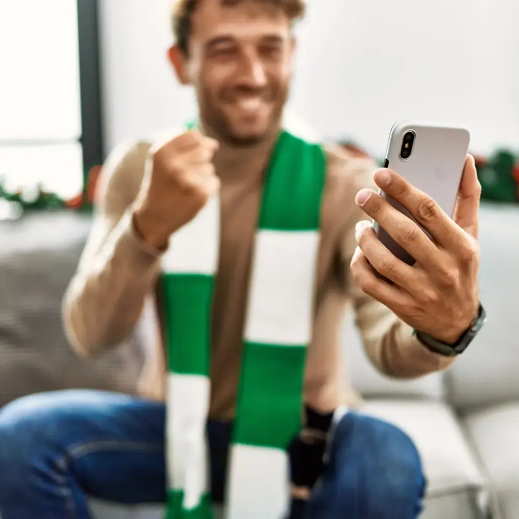 Excited sports fan wearing a green and white scarf celebrates while looking at a smartphone.