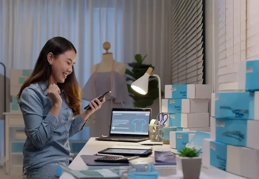 Small business owner celebrating an online sale in a home-based retail workspace with packed orders.