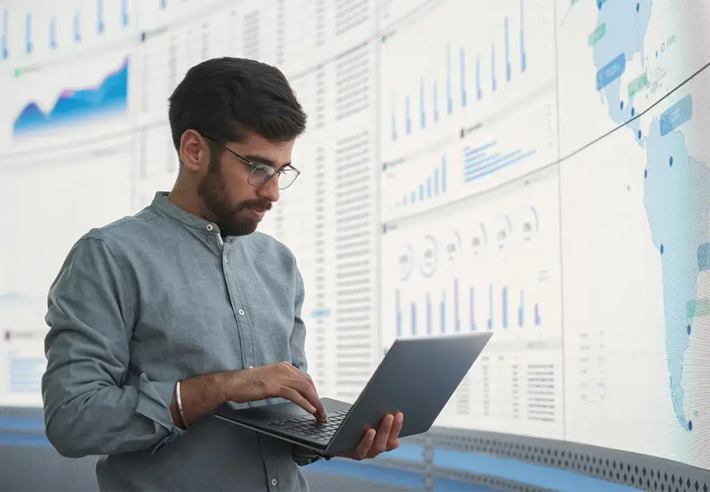 Analyst reviewing global data dashboards on laptop in front of large digital display wall.
