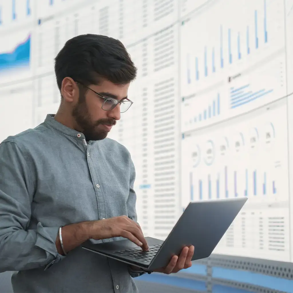 Analyst reviewing global data dashboards on laptop in front of large digital display wall.
