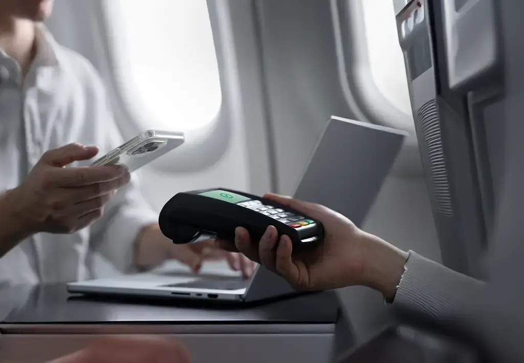 Passenger uses smartphone for contactless payment via card reader on airplane tray table.