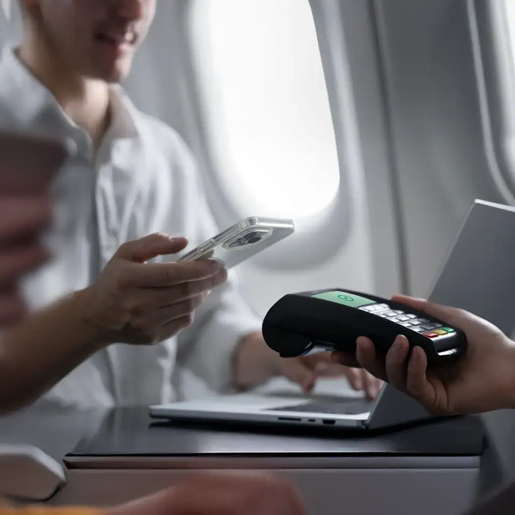 Passenger uses smartphone for contactless payment via card reader on airplane tray table.