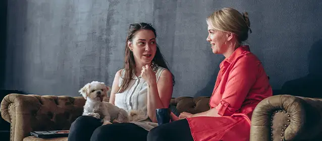 Two colleagues sitting on a sofa in a relaxed office setting, having a conversation with a small dog nearby.