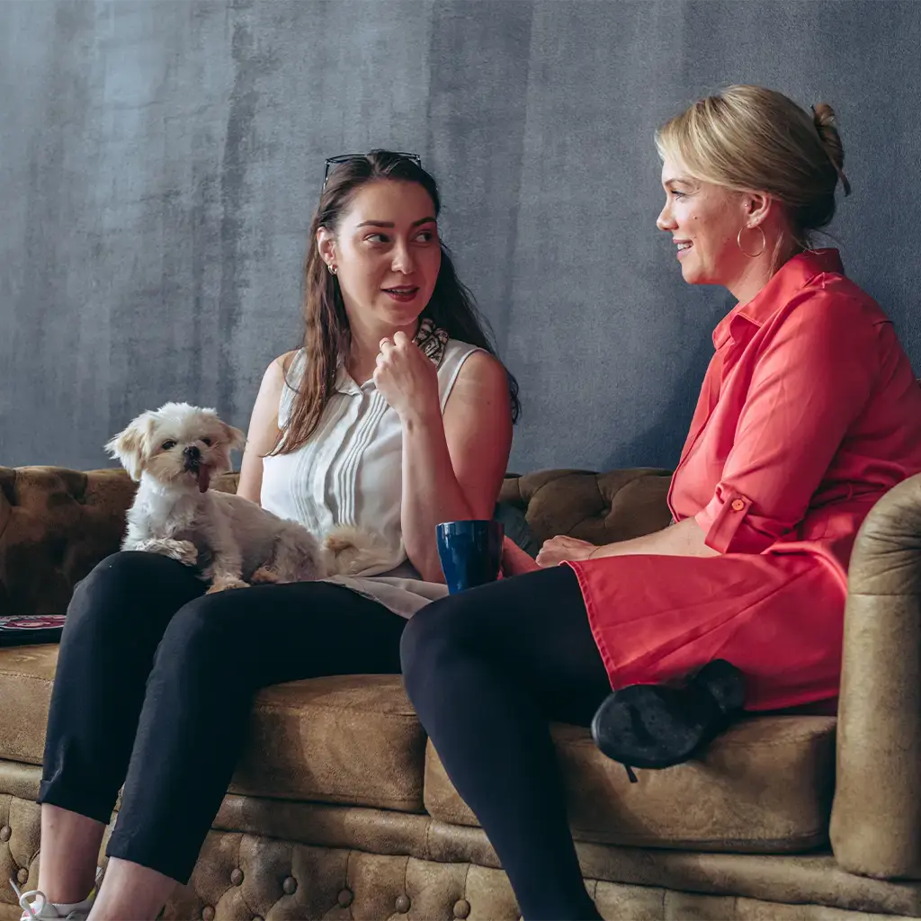 Two colleagues chatting on a lounge sofa in a relaxed office setting, with a small dog beside them