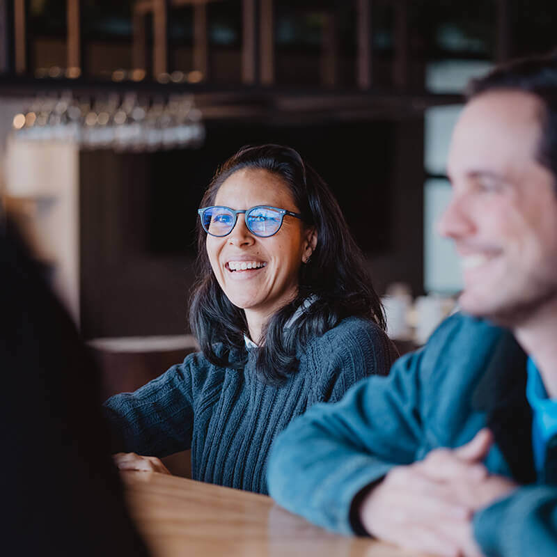 Colleagues smiling and engaged in conversation around a wooden table in a relaxed office environment.