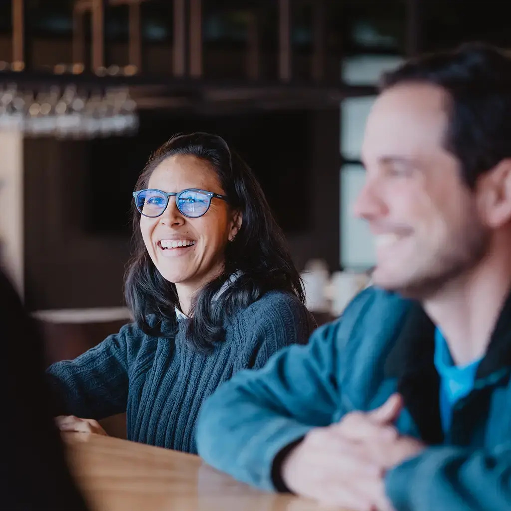 Colleagues smiling and engaged in conversation around a wooden table in a relaxed office environment.