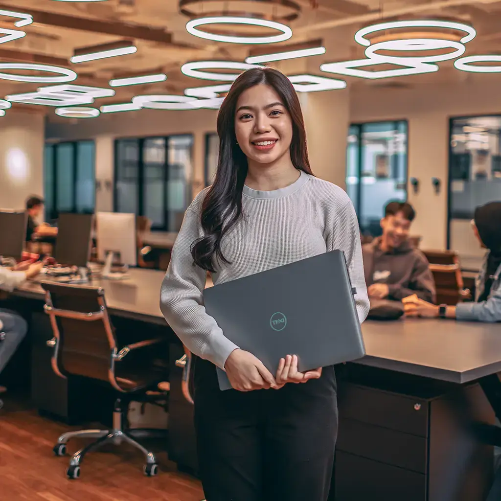 Professional in modern office holding laptop, with team collaborating in background.
