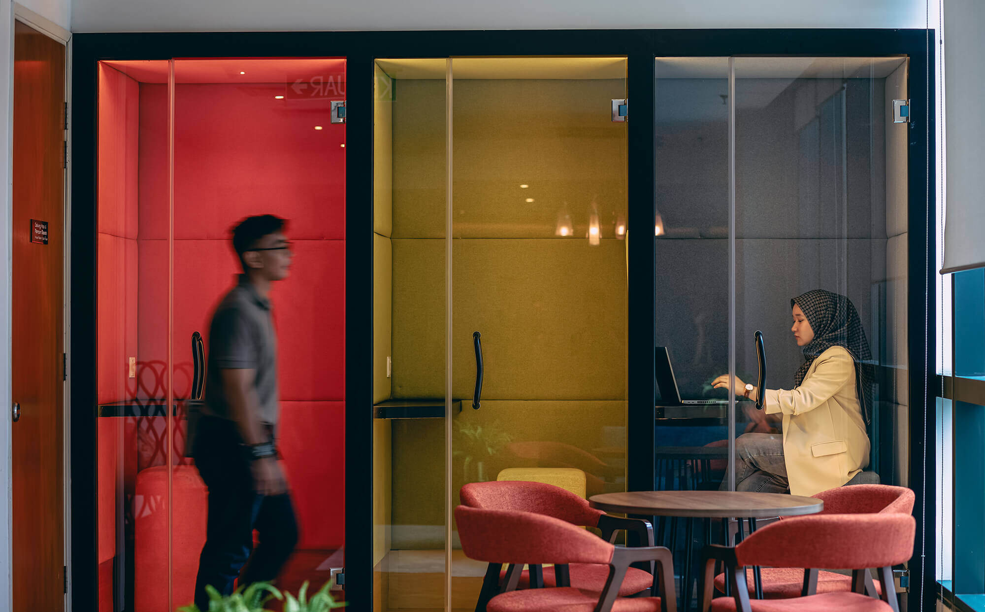 A professional working in a colorful private booth while another walks by in a modern office lounge