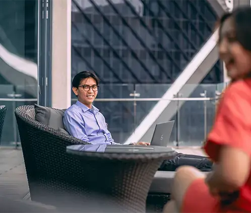 A professional working on a laptop in an outdoor office lounge, engaging in conversation with a colleague.