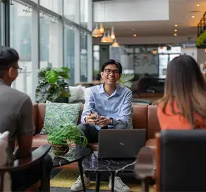 Three colleagues having a discussion in a modern office lounge with greenery and a laptop on the table.