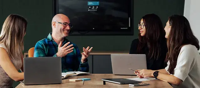 A group of professionals collaborating around a table with laptops and a monitor in a conference room.