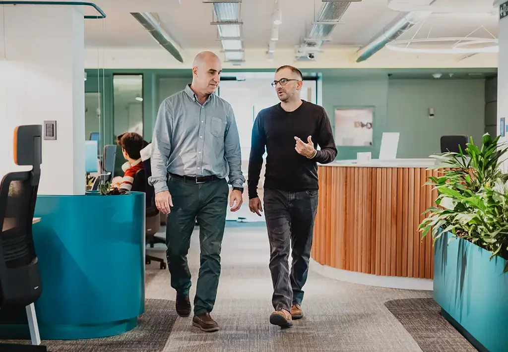 Two colleagues walking and discussing in a modern, open-plan office environment.