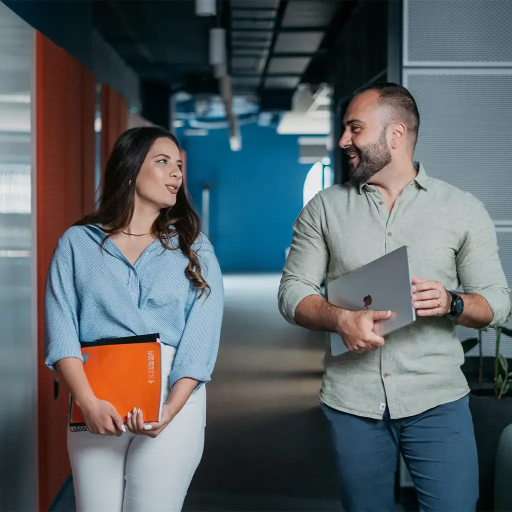 Two colleagues walking through an office corridor while discussing work, carrying a laptop and project folder