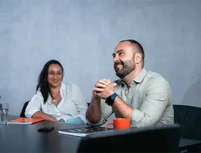 Two colleagues engaged in a discussion during a meeting, seated at a table with notebooks and a coffee cup