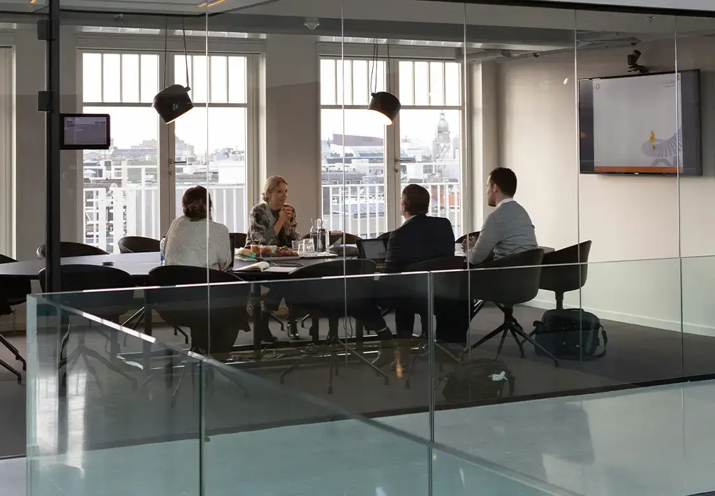 Team in modern glass-walled meeting room discussing strategy with presentation screen in background.