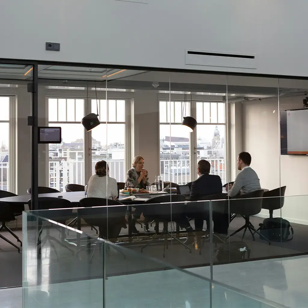 Team in modern glass-walled meeting room discussing strategy with presentation screen in background.