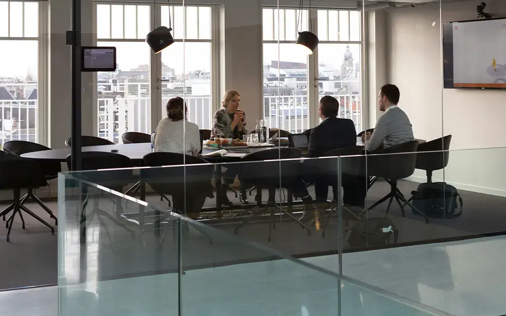 Team in modern glass-walled meeting room discussing strategy with presentation screen in background.