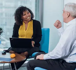 Two professionals engaged in a conversation by a window in a modern office, with a laptop and small table nearby