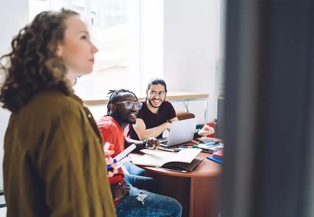 Team collaborating in a modern office, with two colleagues smiling at a laptop while another presents ideas on a whiteboard.