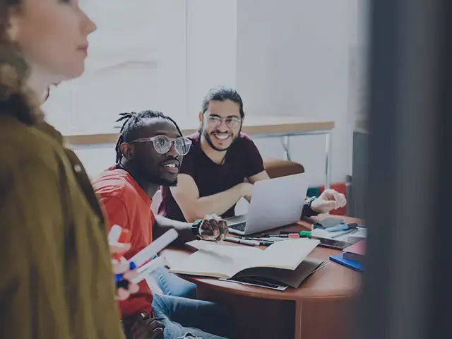 Team collaborating in a modern office, with two colleagues smiling at a laptop while another presents ideas on a whiteboard.