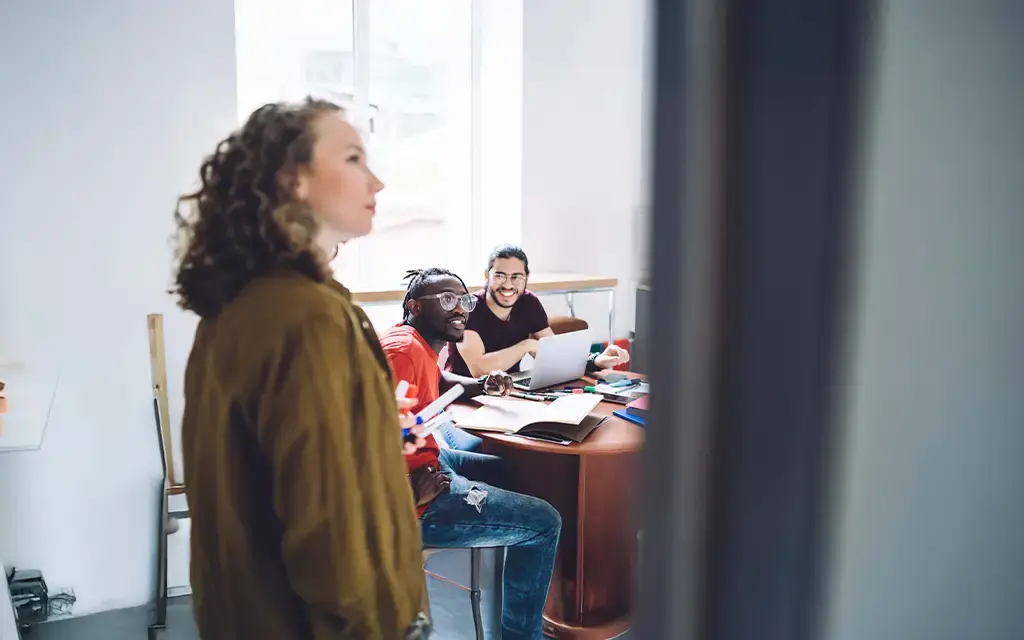 Team collaborating in a modern office, with two colleagues smiling at a laptop while another presents ideas on a whiteboard.