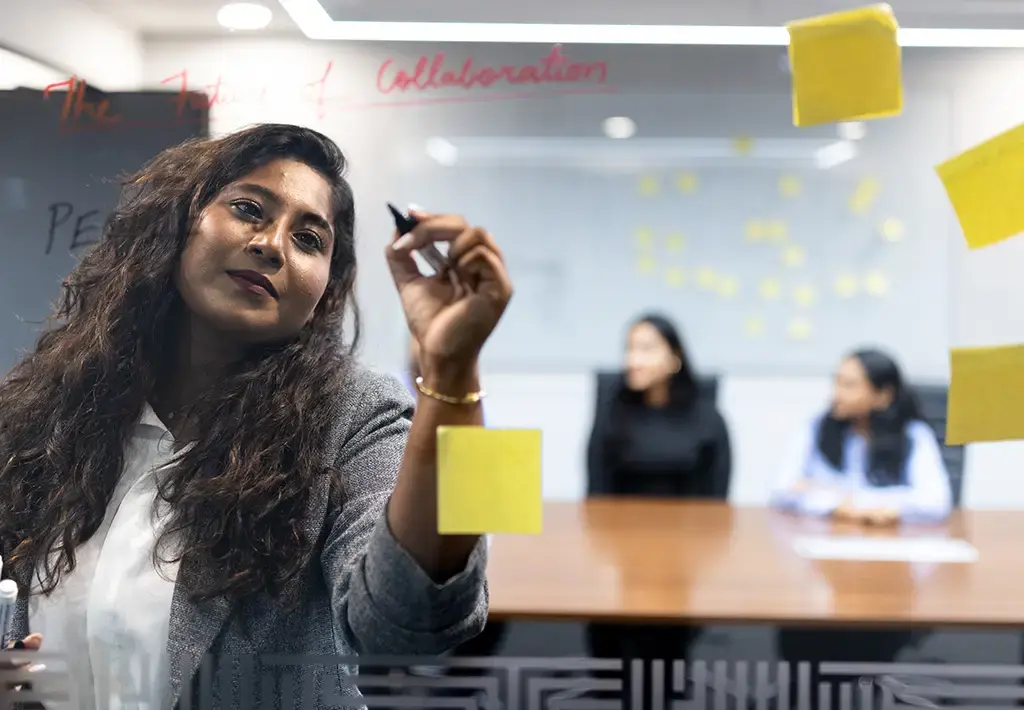 Team member writes on glass wall during a strategy session with sticky notes and colleagues in the background.