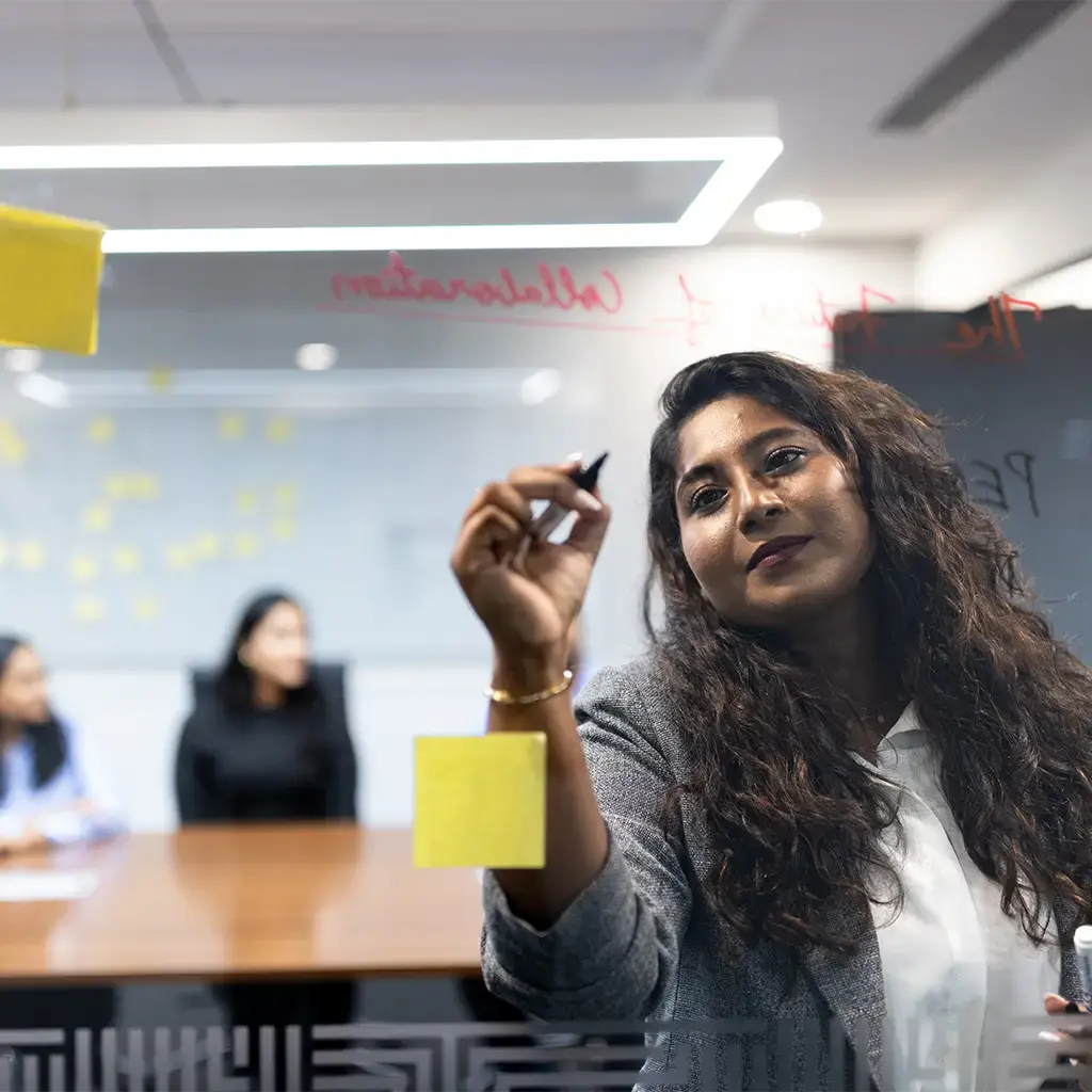 Team member writes on glass wall during a strategy session with sticky notes and colleagues in the background.