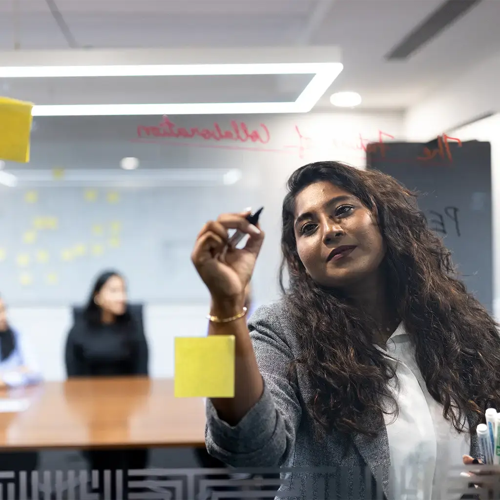Team member writes on glass wall during a strategy session with sticky notes and colleagues in the background.