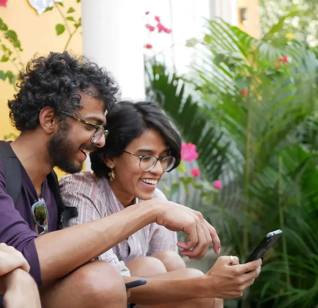 Two people smiling while looking at a smartphone outside near green plants.