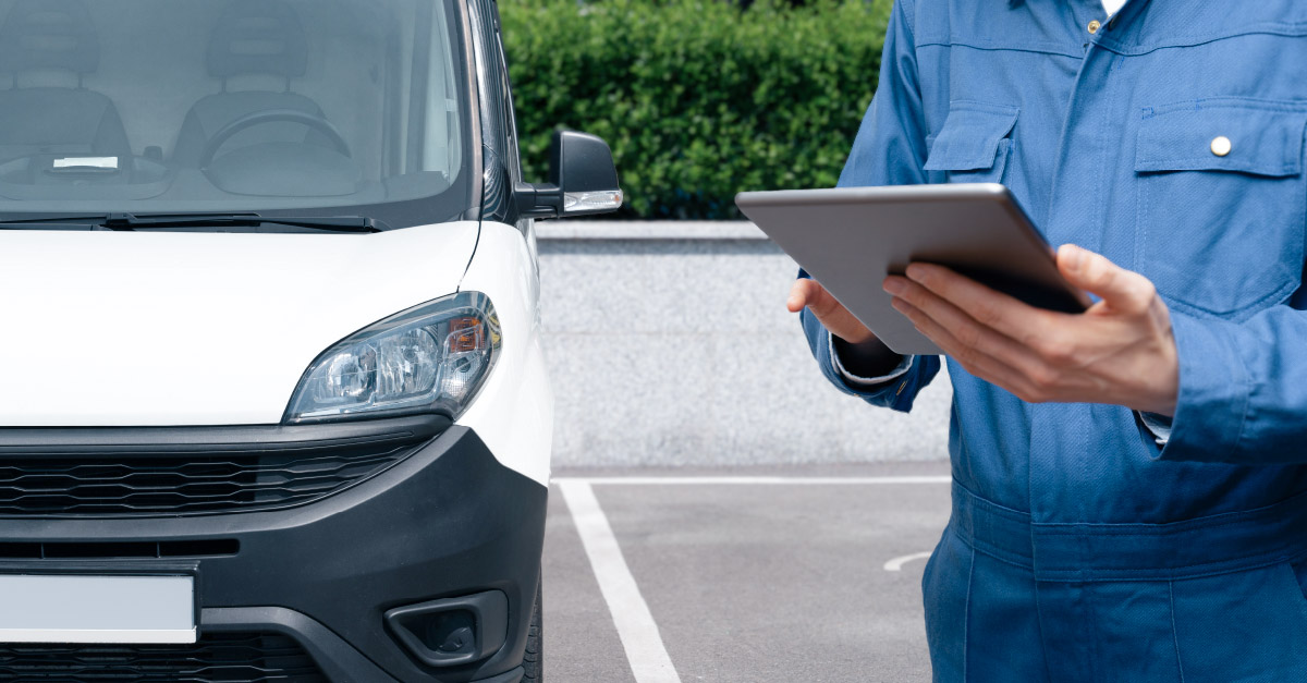 A person in a blue jumpsuit holds a tablet beside a white vehicle with a black front bumper, parked outdoors near greenery and a stone wall.