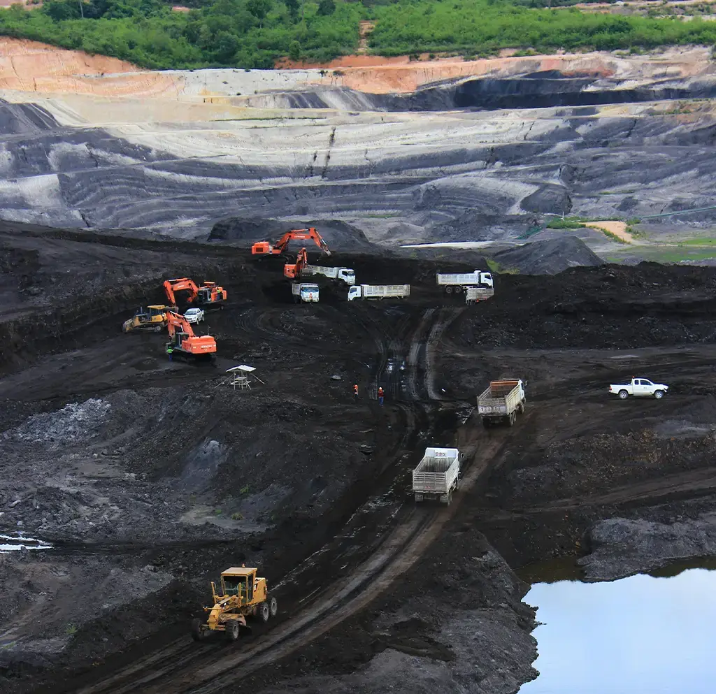 Mining trucks and excavators working in a large open-pit coal mine with layered terrain and water nearby.