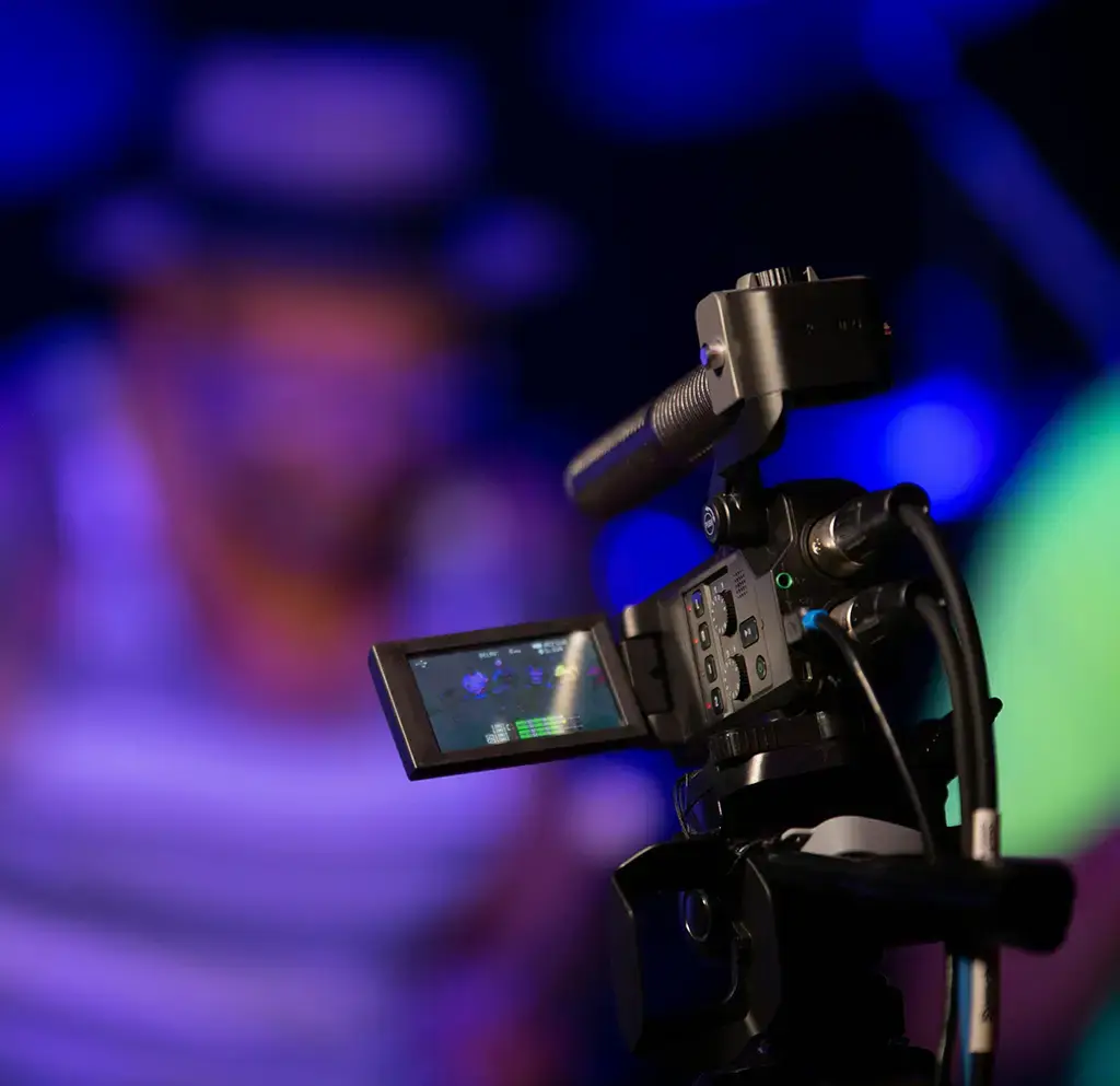 Professional video camera recording a performance in a dimly lit setting with purple lighting.
