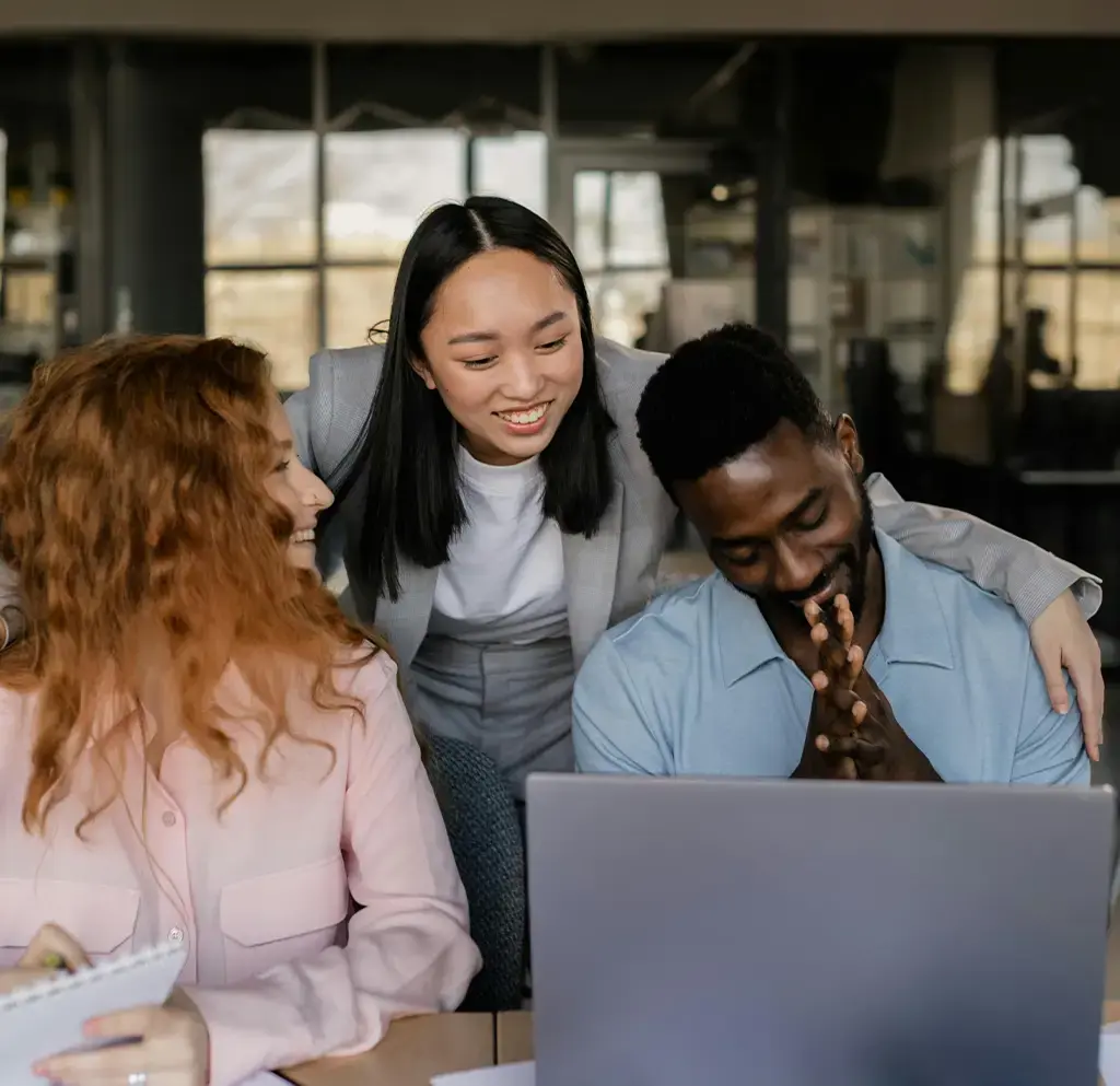Three colleagues collaborating and smiling around a laptop in a modern office with data charts on the table.