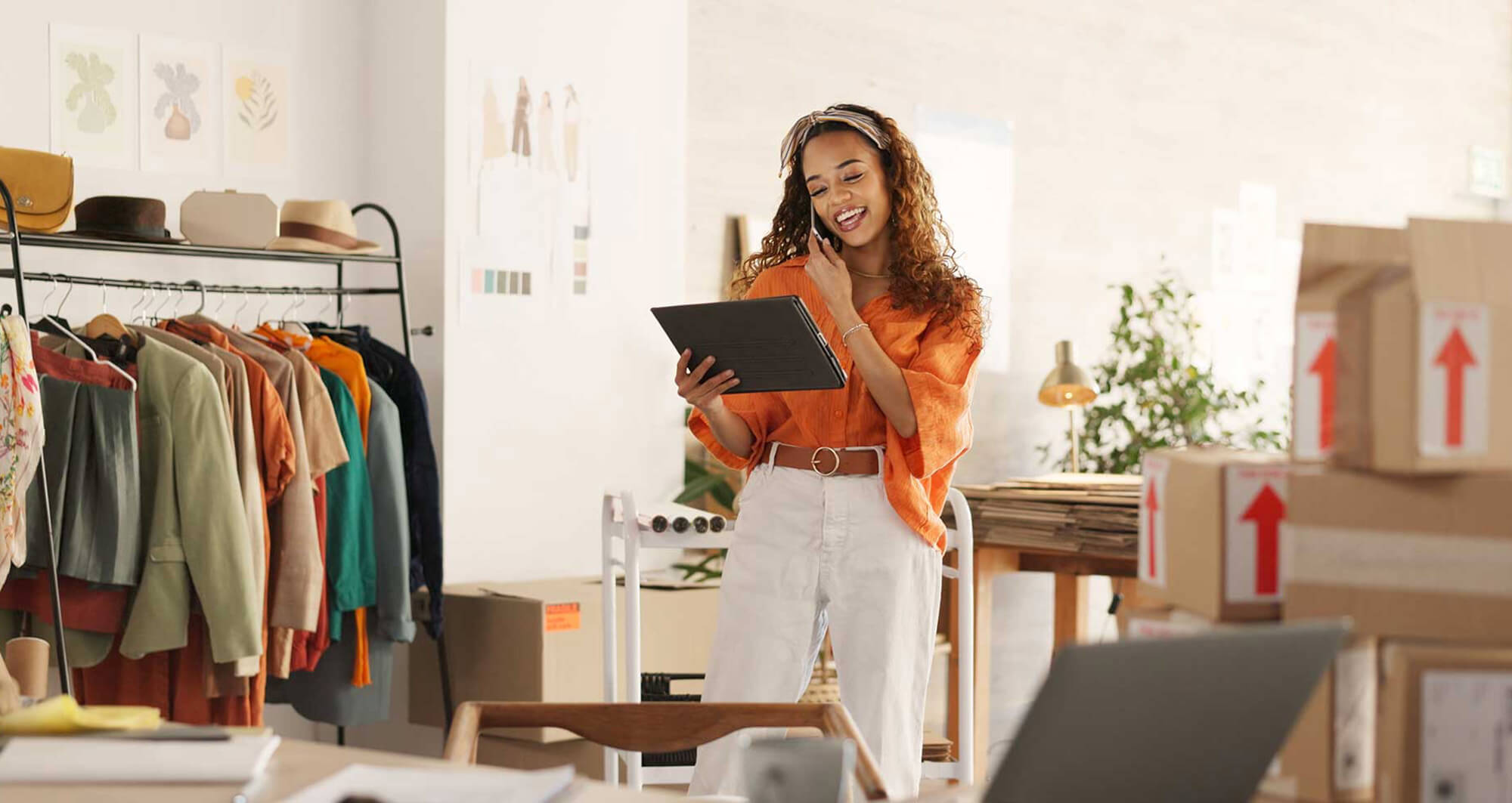 Small business owner using a tablet to manage online fashion retail orders surrounded by clothes and shipping boxes.
