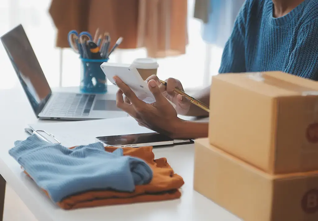 Person managing online orders with clothing, packages, and digital tools at a workspace.