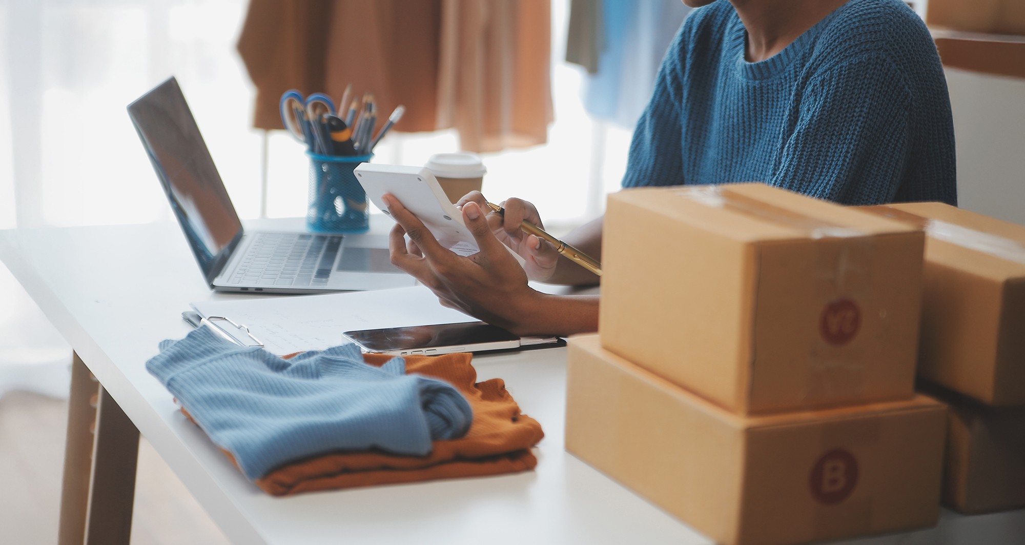 Person managing online orders with clothing, packages, and digital tools at a workspace.