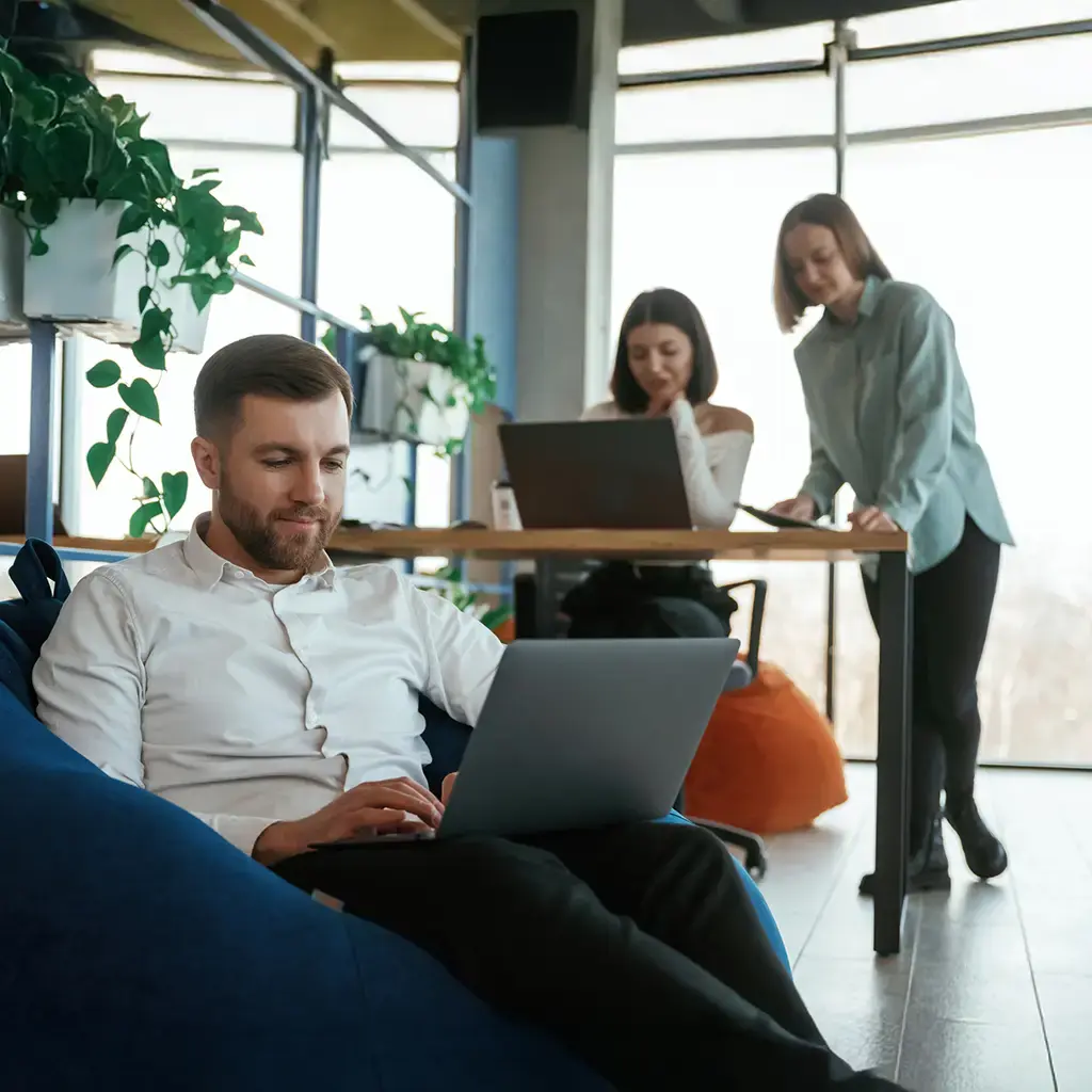 Professionals working on laptops in a relaxed, modern office lounge with natural light and plants.
