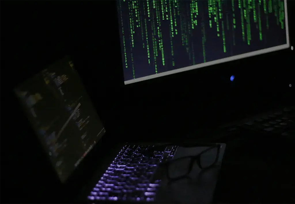 Dark workspace with laptop and monitor displaying code, highlighting cybersecurity or software development.