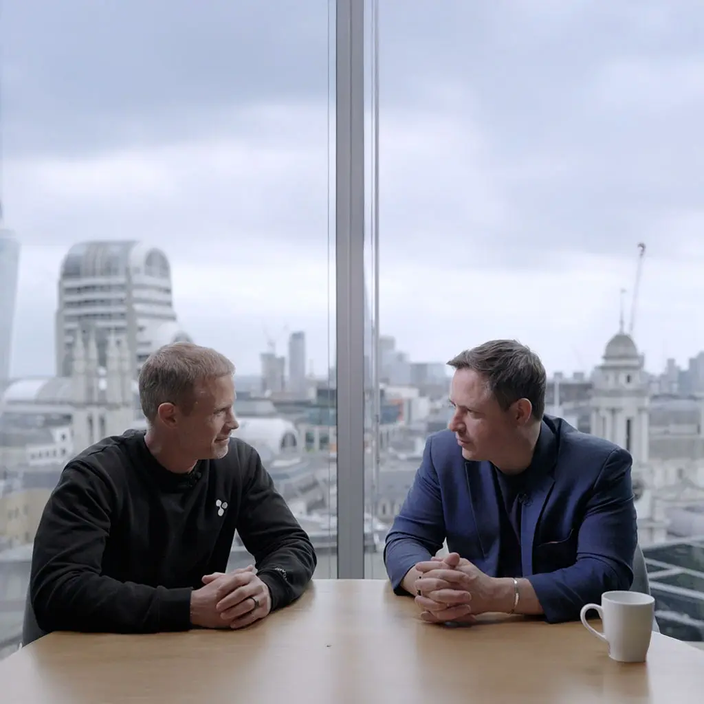 Two business professionals in conversation at a London office with a city skyline view.